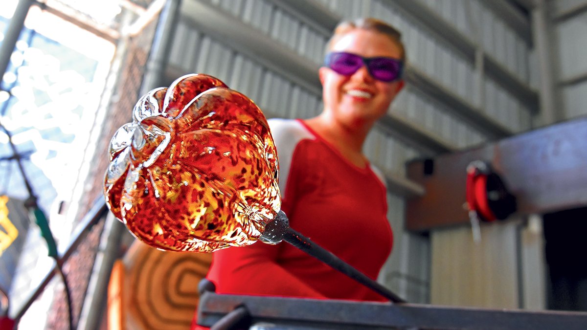 A female glass artist shows off a molten, orange glass art piece in progress at the Morean Arts Center's hot shop in St. Pete