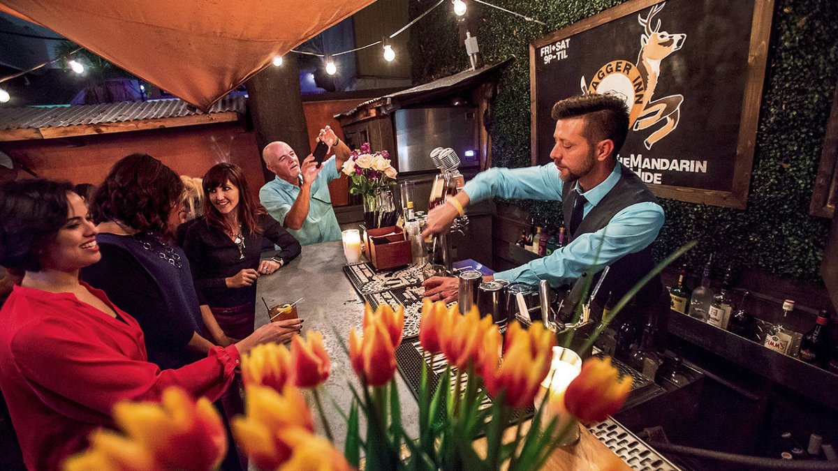 Group of people sitting at a bar with a bartender pouring drinks. Foreground has a vase full of lilies