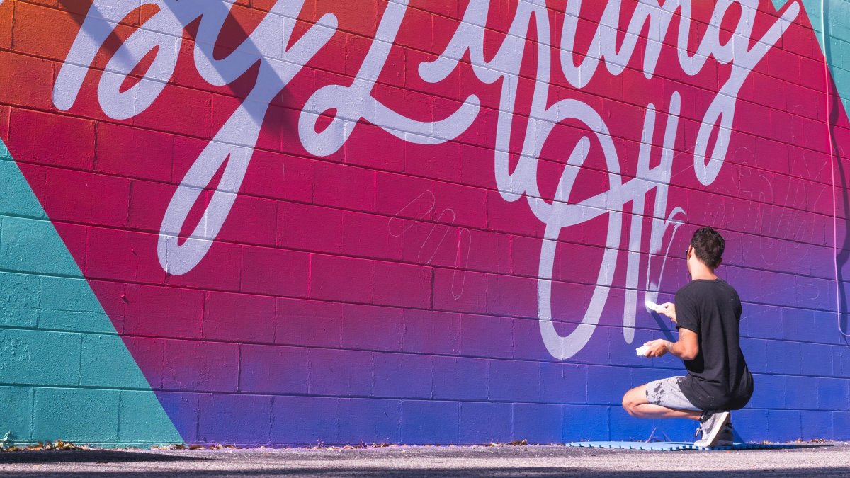 An artist painting a mural on a wall with a teal back ground and gradient from orange to pink to blue with large white writing