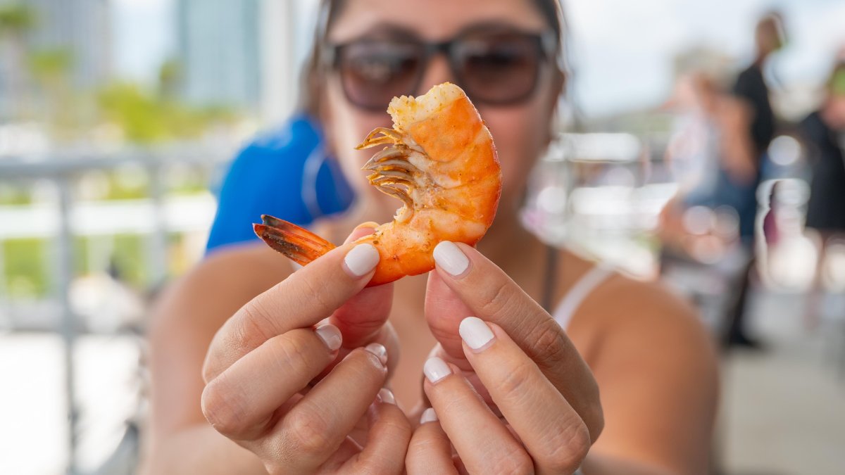 A lady wearing sunglasses holding a shrimp right in front of the camera.