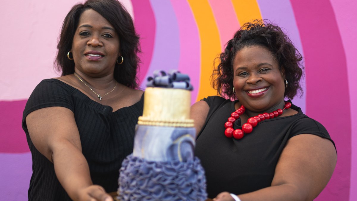 Two women hold up an elaborately decorate cake in front of a bright pink mural