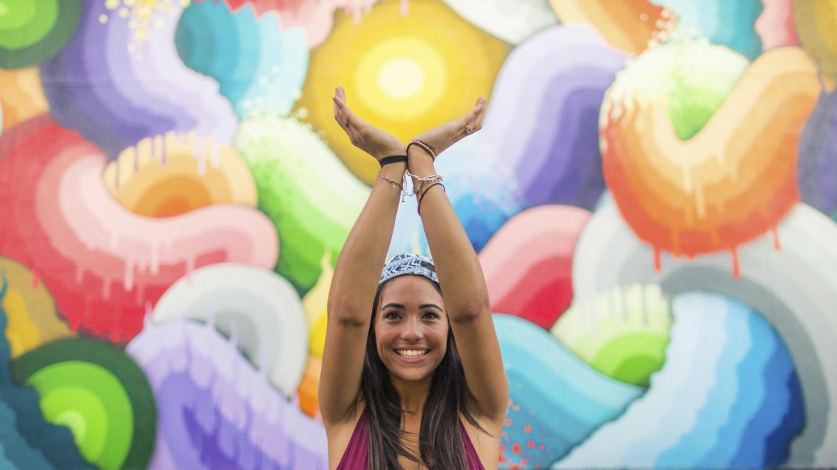 A smiling girl in a red shirt holds her hands up as if to hold the "Eye of the Storm" in a colorful mural by Ricky Watts, Central Arts District