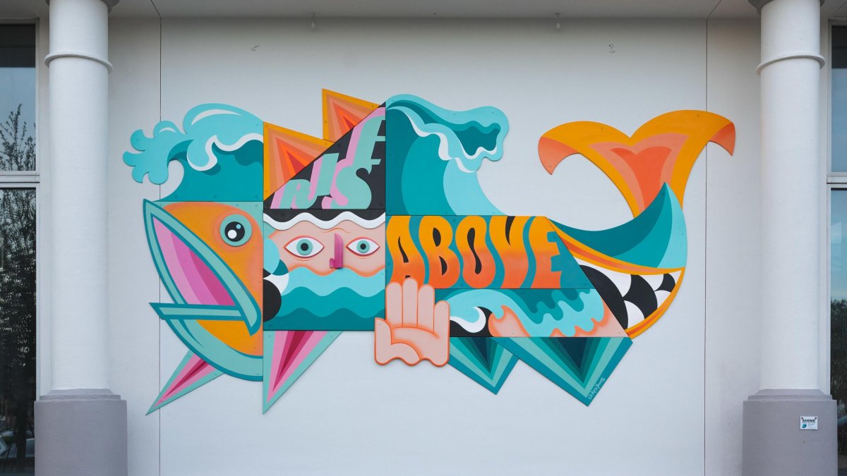 A bright blue, orange and pink mural of a fish created in geometric designs with shapes of a hand and face inside says Rise Above