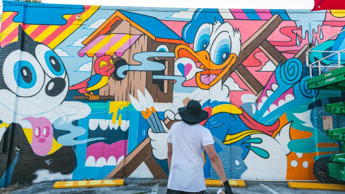 an artist with a spray can of paint in his right hand stands back to look at his work of a cartoonish mural in the EDGE District St. Pete