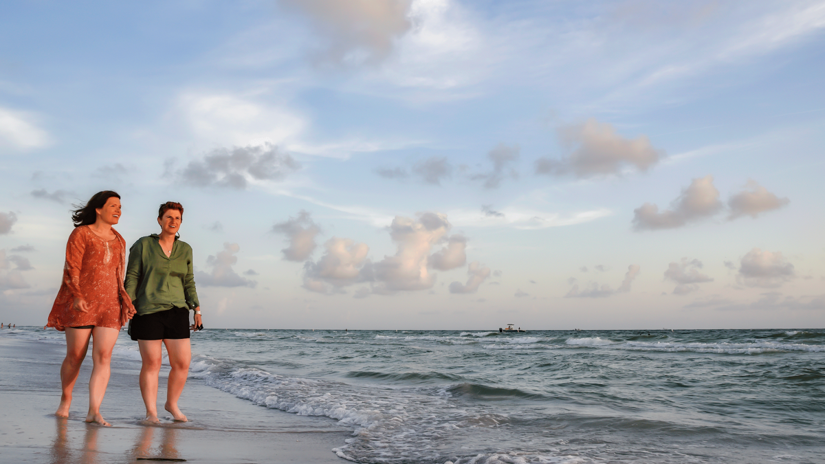 two women hold hands while walking at the water's edge at the beach