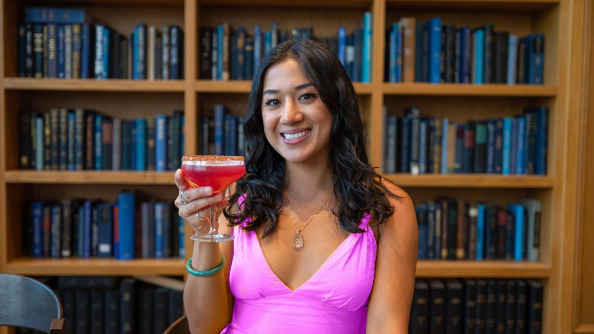 a woman in a Barbie pink dress smiles and holds up a pink cocktail with a background of bookshelves at The Library restaurant