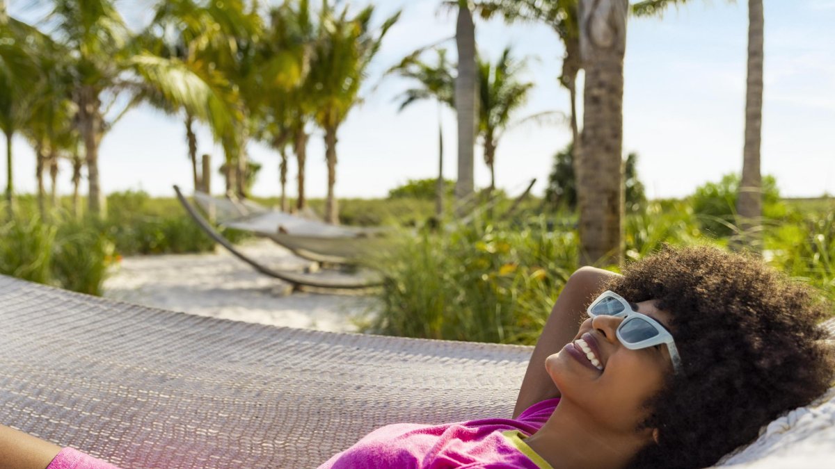 Woman with sunglasses laying in a hammock with the beach and palm trees in the background