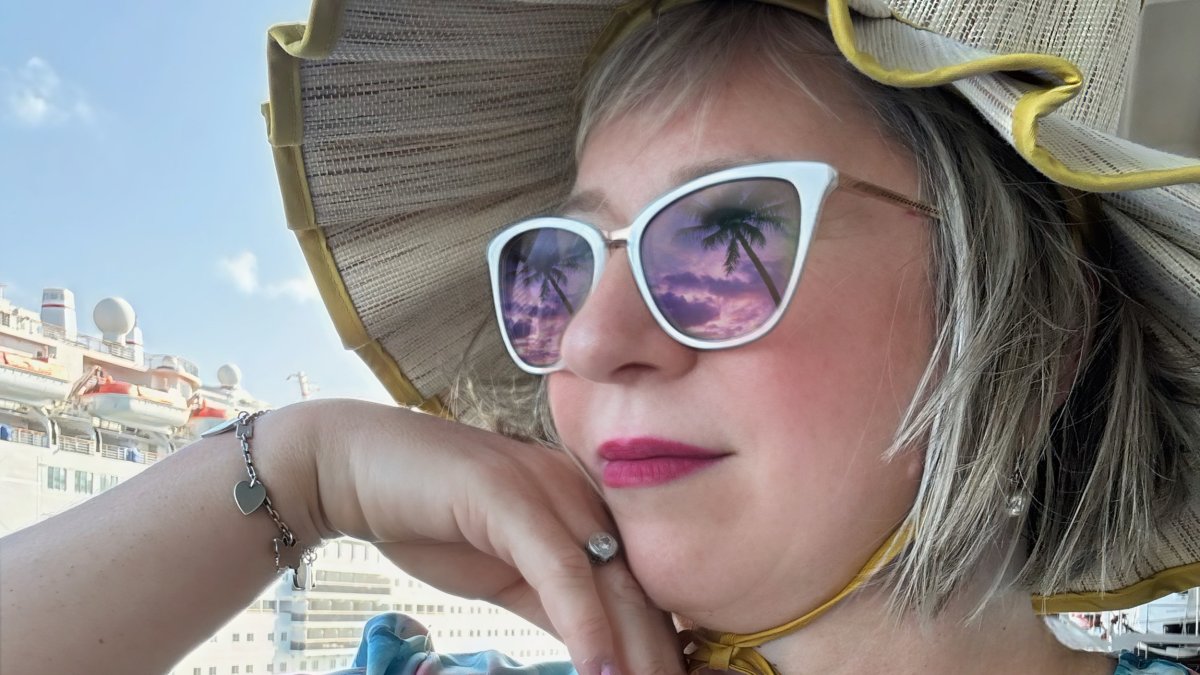 a close-up shot of a woman's face with white sunglasses that are reflecting palm trees and a beach scene; she is wearing a ruffle-edged straw hat and a multi-colored top