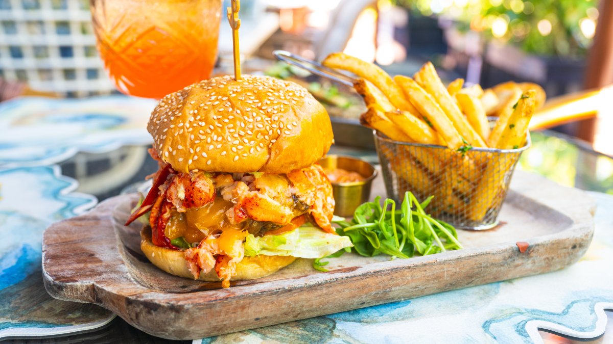 a wooden tray with a burger topped with butter-poached lobster on a sesame seed bun next to a basket of crispy fries at The Helm Seafood Parlour
